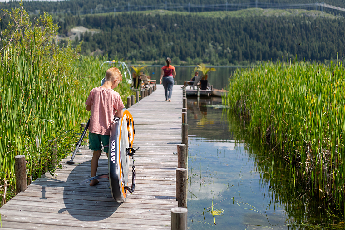 Young boy carrying his paddleboard down a dock surrounded by vibrant green reeds