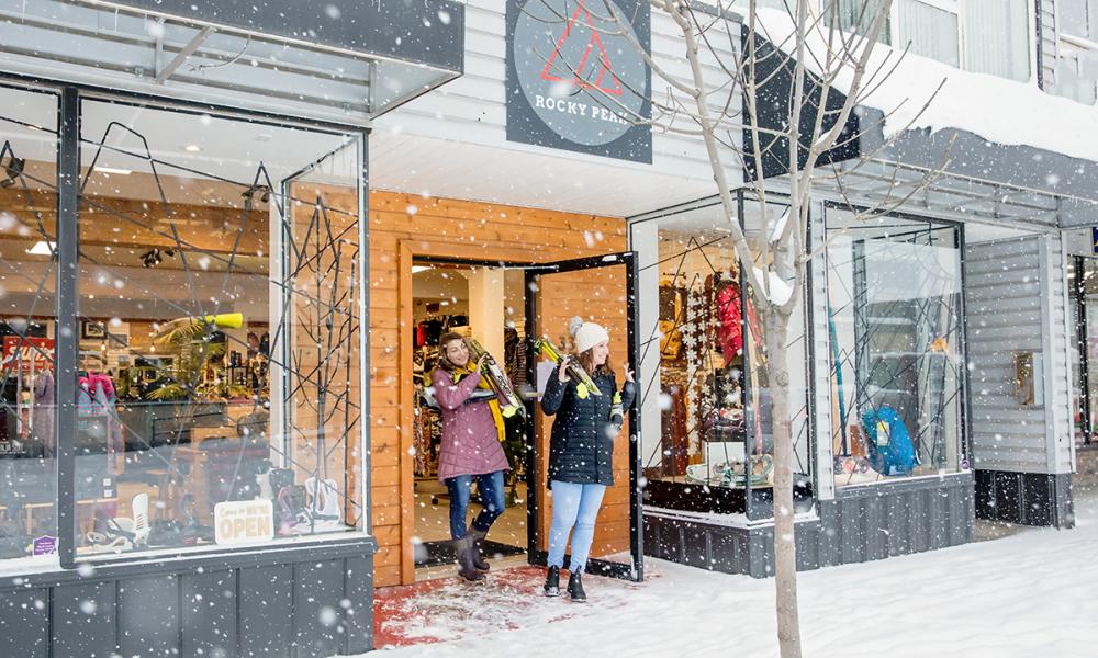Two women exiting an outdoor sports store carrying snowshoes. 