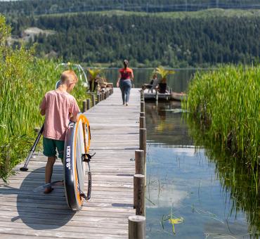 Young boy carrying his paddleboard down a dock surrounded by vibrant green reeds